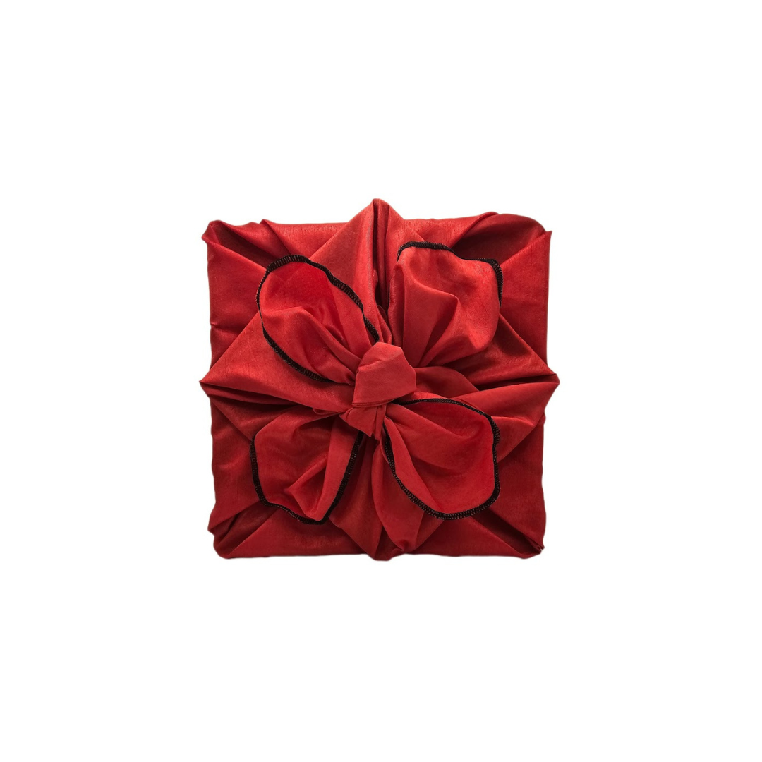 Square gift wrapped in vivid red polyester fabric with a structured decorative knot and contrasting black overlocked edging