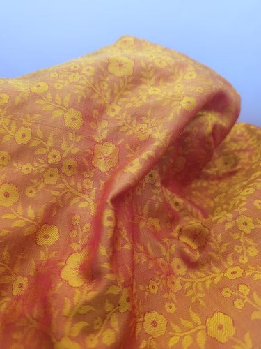 Close-up of orange iridescent silk showing woven flower and leaf motifs, textured surrounding fabric and shifting golden tones