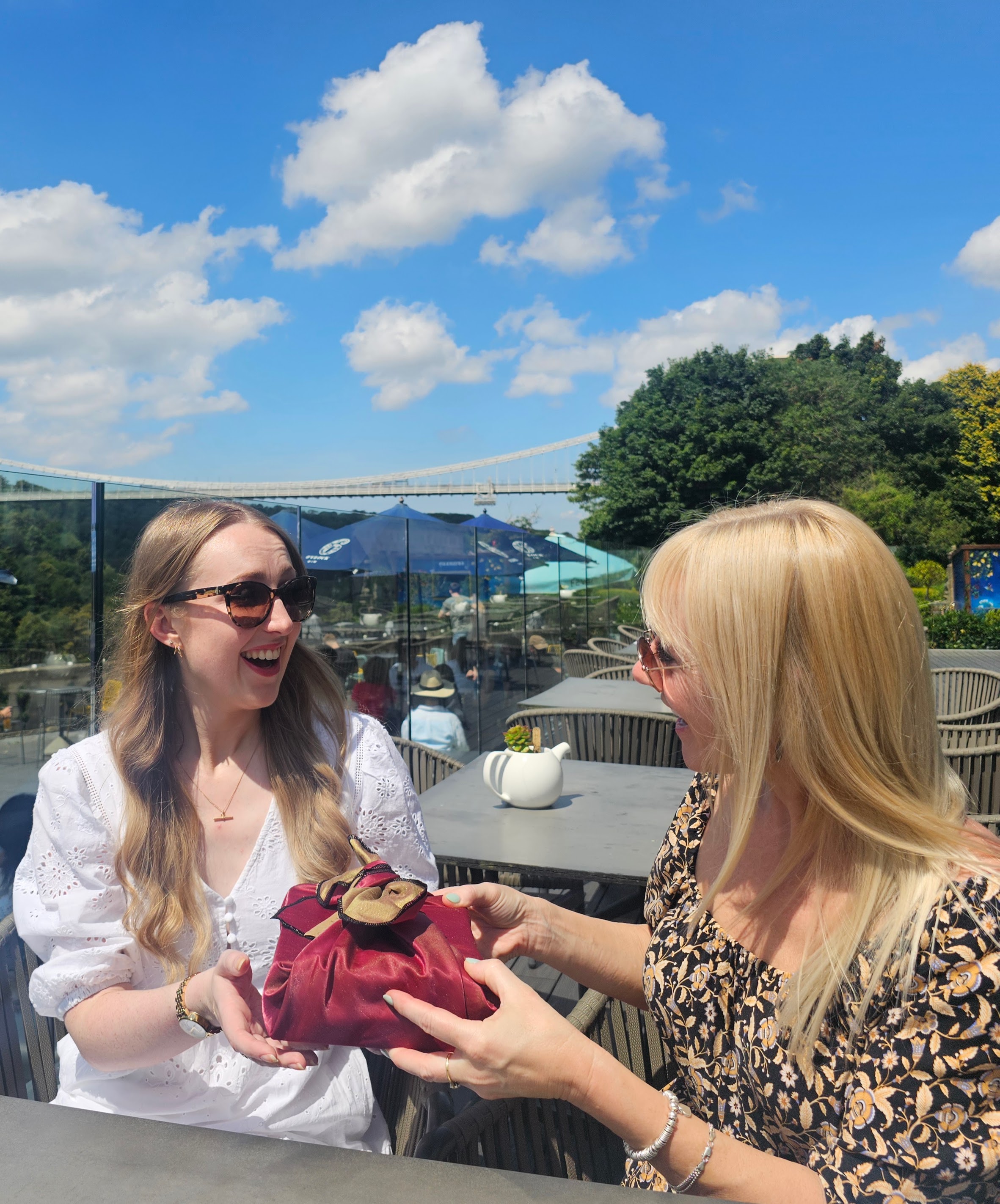 Image of a lady with light blonde hair handing a fabric wrapped gift to a younger lady with strawberry blonde hair