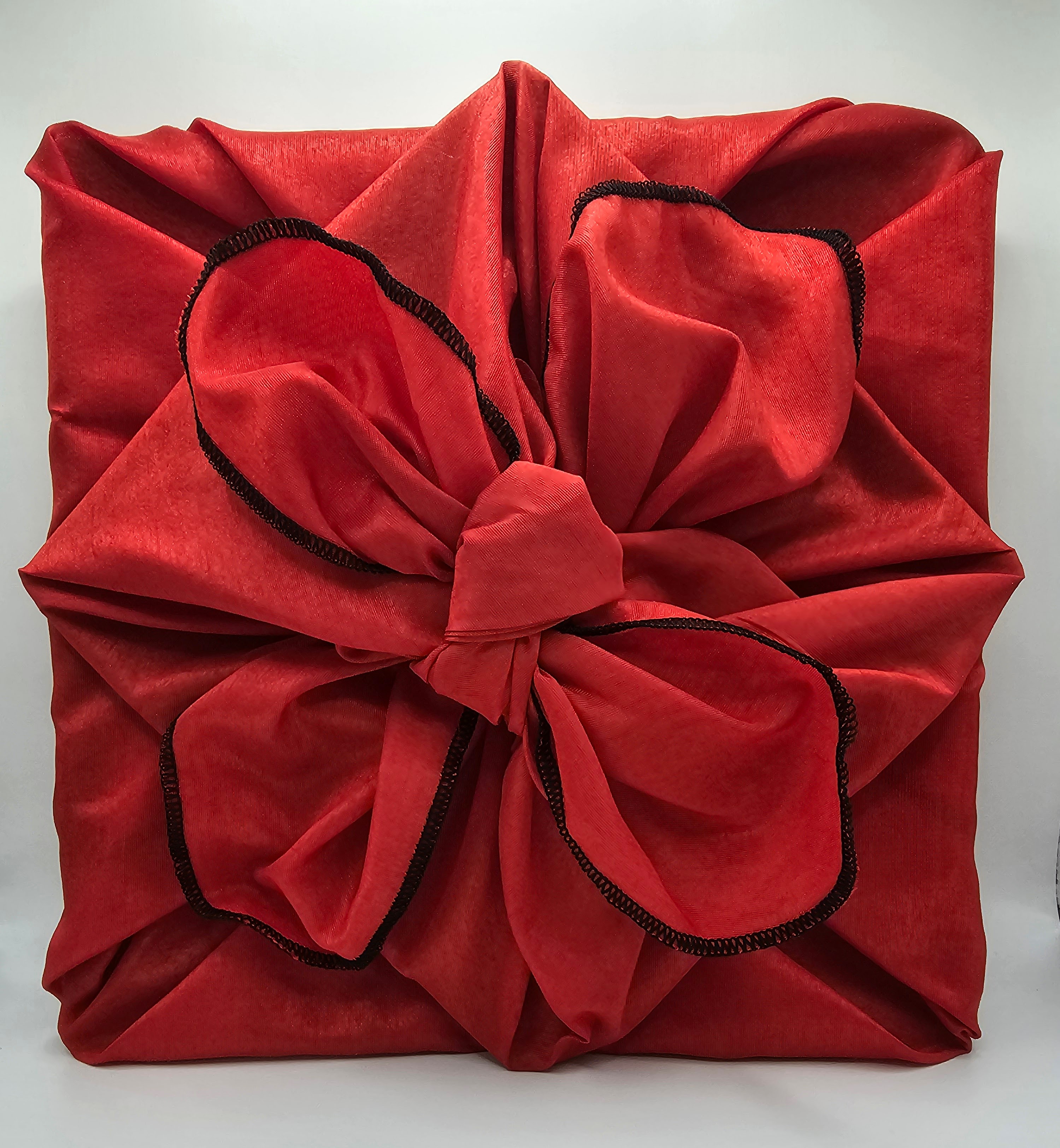 An image showing two red fabric wrapped square flat gifts, the smaller one on top of the larger, placed diagonally with the bow tied to look like the petals of a flower