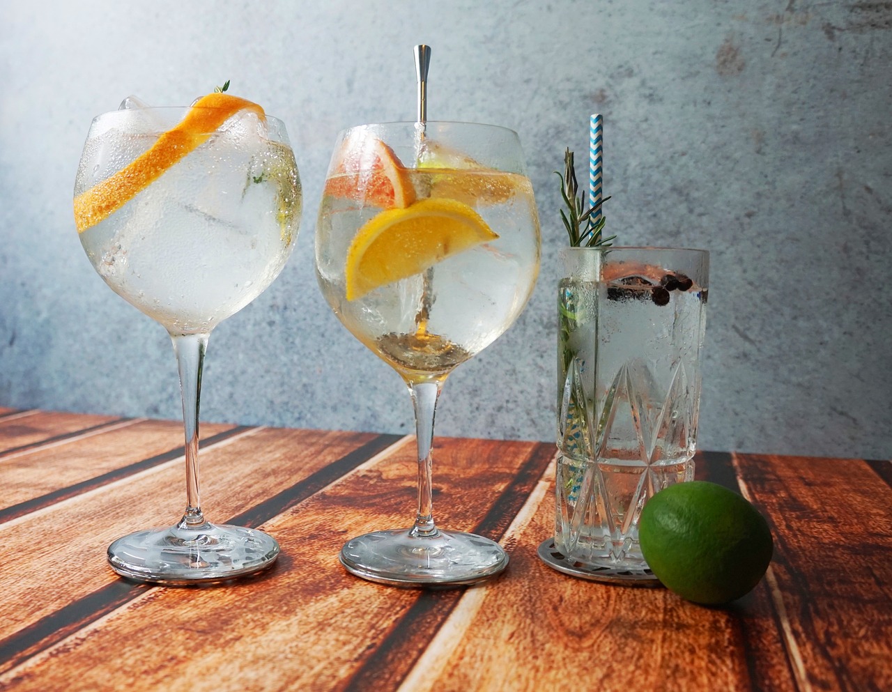 An image showing one straight glass and two different size balloon glasses containing gin, tonic, orange and lemon slices, herbs and straws