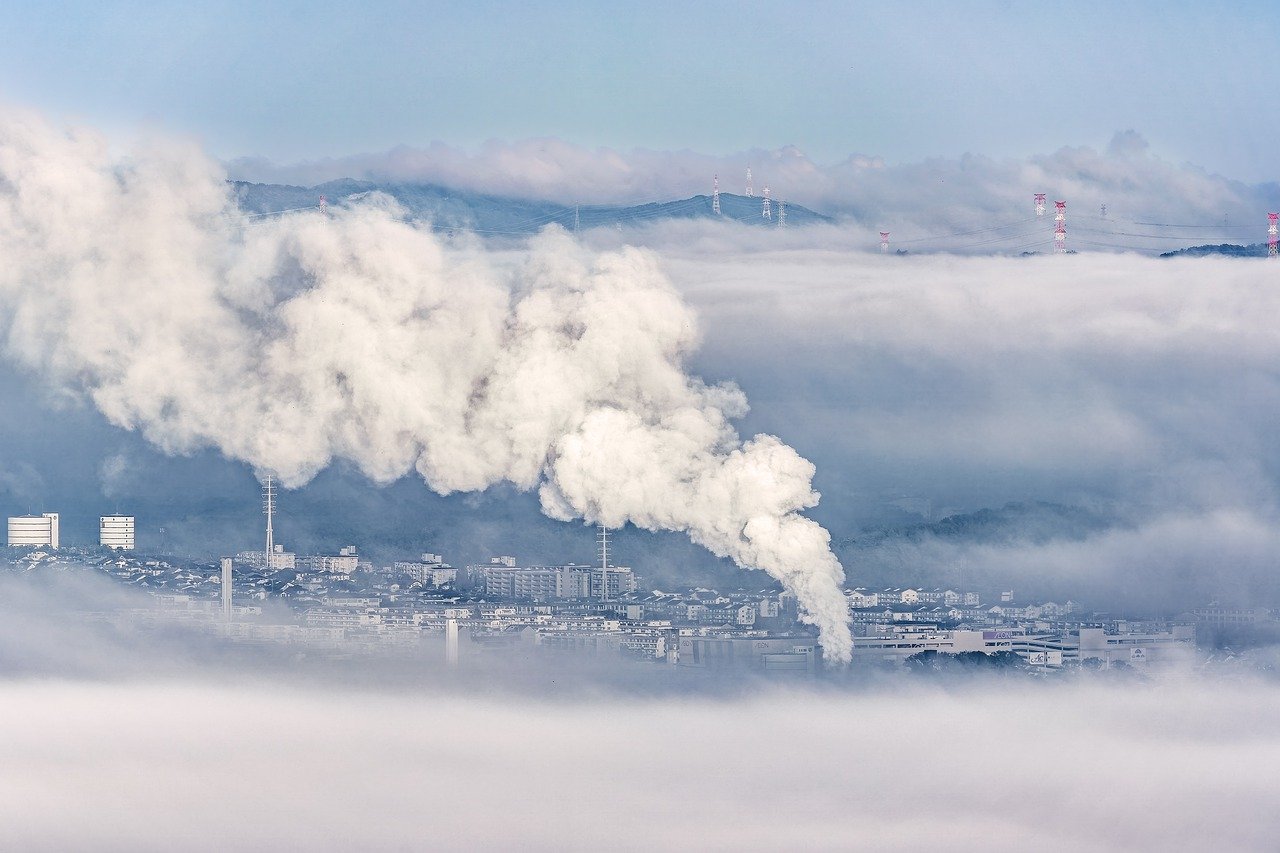 An image of an incinerator in valley in the distance, amongst a highly industrialised area, belching out a big plume of smoke, surrounded by smog and dull blue sky